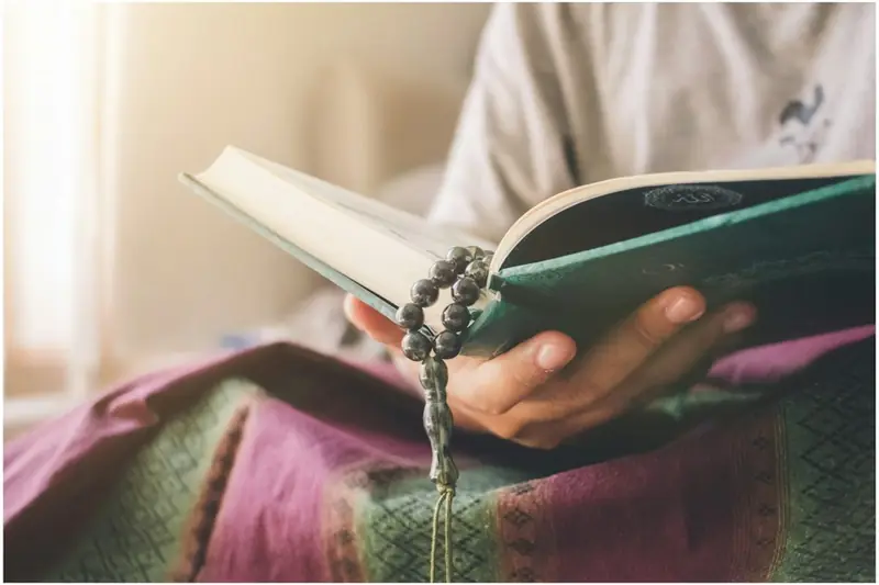 Hands holding an open Quran with prayer beads